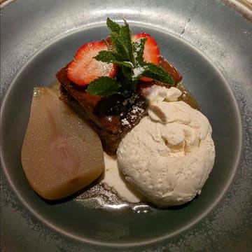 A close-up, overhead shot of a plated dessert featuring a slice of cake, a scoop of vanilla ice cream, a poached pear, and a strawberry garnish.