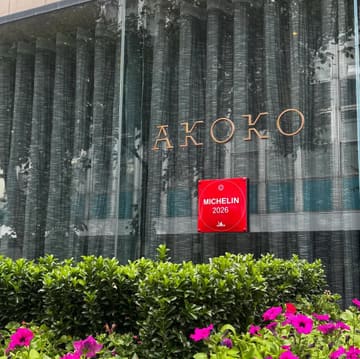 A clean, professional eye-level shot of the exterior of Akoko restaurant, featuring the brand signage and a Michelin 2026 plaque, framed by greenery in the foreground.