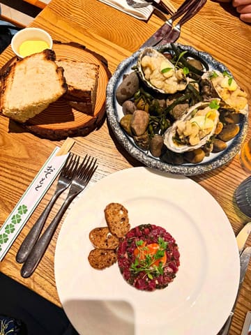 A high-quality overhead shot of a meal featuring beef tartare and tempura mussels, served with bread and butter. The presentation is clean and professional, highlighting the quality of the ingredients and the plating style of the venue.