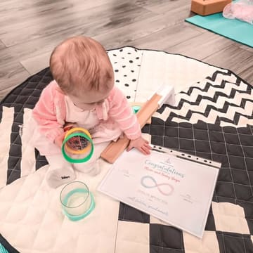 A high-quality, well-composed photograph featuring a baby sitting on a patterned yoga mat, holding a toy, next to a 'Mum and Baby Yoga' certificate. The image is bright, clean, and effectively communicates the calm, welcoming nature of the class.
