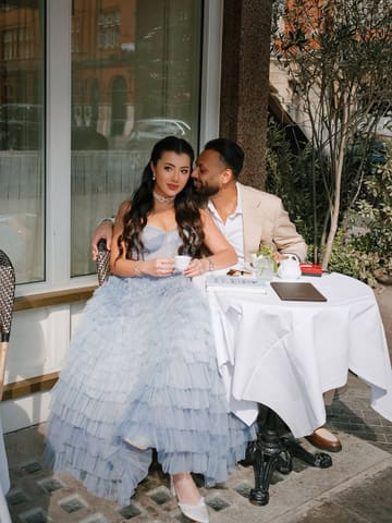 A professionally shot, editorial-style photograph of a couple enjoying coffee at an outdoor cafe table. The image features high-quality lighting and composition, capturing a romantic, aspirational atmosphere suitable for a premium hospitality venue.