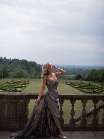 A high-quality, atmospheric shot of a woman in an elegant evening gown standing on the terrace of a historic estate, likely Cliveden House. The image captures a sense of luxury and occasion, focusing on the grandeur of the setting and the fashion.