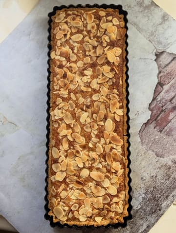 A top-down shot of a homemade gluten-free Bakewell tart, featuring a frangipane filling topped with slivered almonds, presented in a rectangular tart tin on a textured surface.