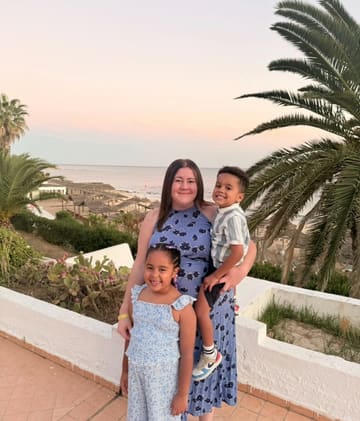A warm, candid family portrait taken at a resort or hotel during sunset. The image captures a mother and her two children posing against a backdrop of palm trees and a beach, effectively conveying a relaxed, holiday atmosphere suitable for travel or hospitality marketing.