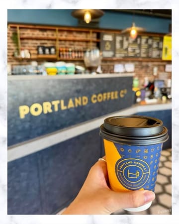 A first-person perspective shot of a hand holding a takeaway coffee cup in front of a coffee shop counter.
