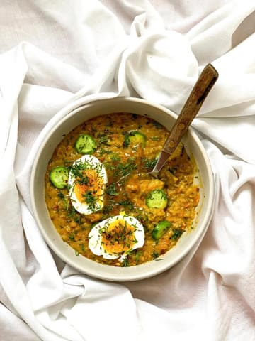 A bright, high-key overhead shot of a bowl of lentil soup garnished with halved boiled eggs, fresh dill, and cucumber slices, presented on a white fabric surface.