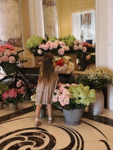 A high-quality, aesthetic lifestyle shot of a child exploring the floral displays in the lobby of The Langham, London. The image captures a sense of luxury and family-friendliness, serving as an inviting glimpse into the venue's atmosphere.