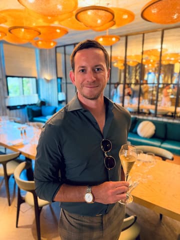 A portrait of a man holding a glass of sparkling wine in an upscale, warmly lit restaurant setting. The image captures a sophisticated atmosphere, likely during a business or social dinner event.
