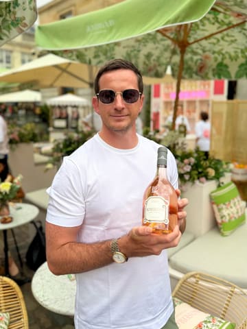 A portrait of a man holding a bottle of Roseblood d’Estoublon wine, set against the backdrop of a floral-decorated outdoor terrace in Covent Garden.