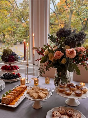 A beautifully styled afternoon tea spread featuring tiered stands of pastries, scones, and sandwiches, set against a window with soft, warm autumnal light.
