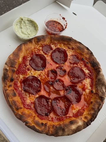 A top-down, overhead shot of a homemade pepperoni pizza served in a cardboard delivery box, accompanied by two small plastic tubs of sauce.