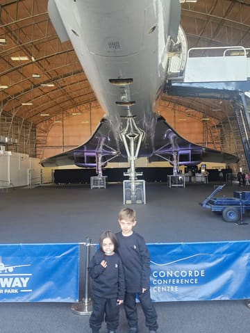 A photo of two children standing in front of a Concorde aircraft inside a large hangar, likely at a museum or aviation attraction.