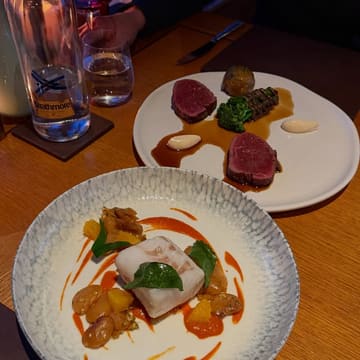 A high-angle, close-up shot of two plates of food on a wooden table in a dimly lit restaurant setting. The foreground plate features a piece of white fish with a vibrant orange sauce and beans, while the background plate displays two pieces of meat with broccoli and sauce dots.