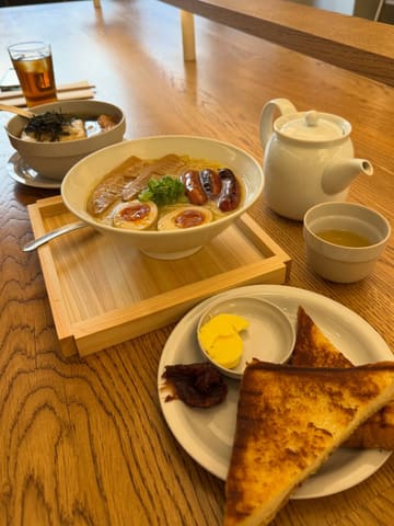 A high-angle, well-composed shot of a Japanese breakfast spread featuring ramen with sausage, udon with fried tofu, and toast with butter and red bean paste, set on a wooden table.