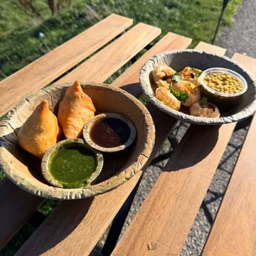 A photo of Indian street food, specifically samosas with chutneys and pani puri, served in disposable bowls on a wooden table at an outdoor beachfront location.