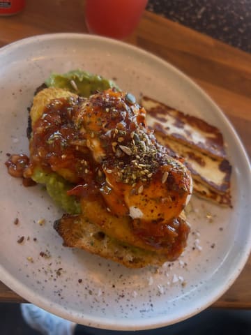 A top-down, close-up shot of a brunch dish featuring poached eggs, avocado, hash browns, and halloumi on toast, served on a speckled plate.