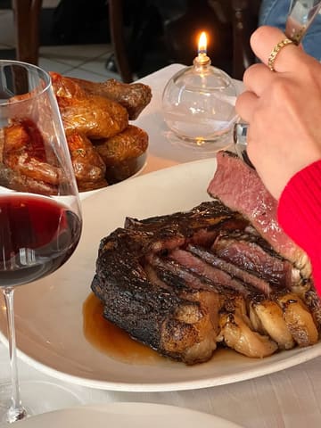 A close-up, atmospheric shot of a bone-in sirloin steak being served at a restaurant, accompanied by a glass of red wine and a side of potatoes. The lighting is warm and intimate, typical of a high-end dining environment.