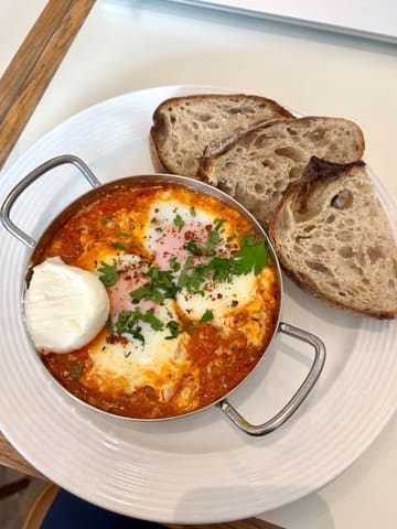 A clean, top-down shot of a shakshuka dish served in a metal pan, accompanied by slices of sourdough bread on a white table. The lighting is bright and natural, highlighting the textures of the eggs and sauce.