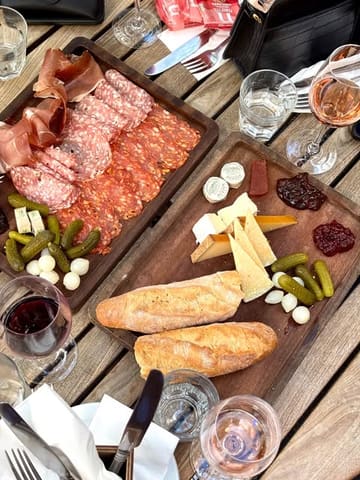 A high-quality, overhead flat-lay shot of two wooden sharing boards featuring charcuterie, various cheeses, crusty bread, pickles, and wine, set on a rustic wooden table.
