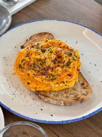 A high-quality, well-lit close-up shot of scrambled eggs on sourdough toast, likely taken at a brunch venue. The composition is clean and the food is presented in an appealing, appetizing manner.