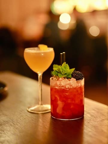 A high-quality, atmospheric shot of two cocktails on a wooden bar top, capturing the moody, upscale vibe of a cocktail bar.
