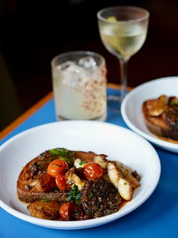 A high-quality, professional-looking food photograph featuring a dish of cod cheeks, chorizo, tomato, and morcilla on sourdough, accompanied by a cocktail. The image is well-composed with a shallow depth of field, highlighting the textures and colours of the food.