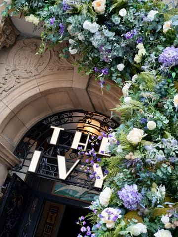 A high-quality, eye-level shot of the entrance to The Ivy Glasgow, featuring a lush, vibrant floral archway framing the restaurant's signage. The composition is clean and symmetrical, highlighting the elegant architectural details of the building facade.