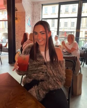 A portrait of a woman holding a cocktail in a casual bar or restaurant setting. The image captures a social moment, though the lighting is inconsistent with a distracting warm light spill on the subject's face.