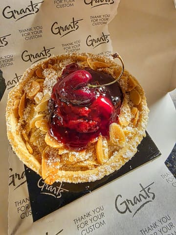A close-up, overhead shot of a cherry frangipane tart presented inside a branded bakery box. The image highlights the textures of the pastry, almond topping, and the glossy cherry glaze, though the overall aesthetic is impacted by the use of direct, harsh flash photography.