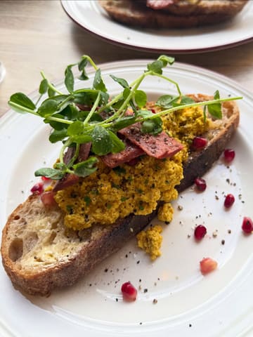 A close-up, top-down shot of a savoury brunch dish featuring scrambled eggs (likely tofu-based) on a slice of sourdough toast, topped with vegan bacon strips and pea shoots, served on a white plate.