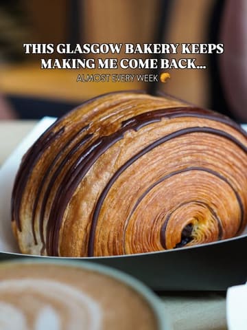 A high-quality, close-up shot of a chocolate-striped pastry, likely a pain au chocolat, served in a paper tray. The image uses a shallow depth of field to blur a coffee cup in the foreground, creating a professional, inviting aesthetic typical of high-end bakery marketing.
