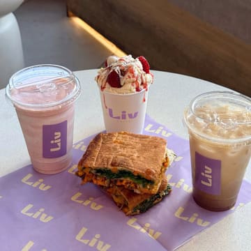 A clean, well-composed, and highly appetising shot of a cafe spread featuring a smoothie, a toasted sandwich, and a dessert cup, all branded with the 'Liv' logo. The lighting is soft and natural, effectively highlighting the textures of the food, while the branded paper liner provides a cohesive and professional look that is perfectly suited for social media marketing.