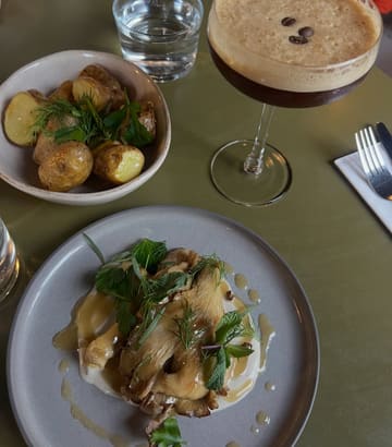A high-quality, overhead food photography shot featuring a vegan lunch spread consisting of BBQ mushrooms with cashew cream, charred potatoes, and an espresso martini. The lighting is soft and atmospheric, typical of a modern restaurant setting, and the composition is clean and well-balanced.