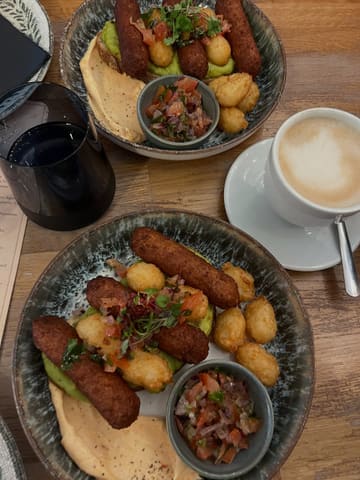 A top-down, casual photograph of two brunch plates featuring veggie sausages, smashed avocado on sourdough, hash browns, and salsa, accompanied by a coffee. The image captures a typical cafe dining experience, focusing on the meal rather than the environment.