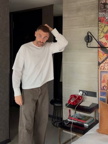 A casual, lifestyle-oriented shot of a man standing in a hotel room, likely part of a 'day in the life' or travel content series.