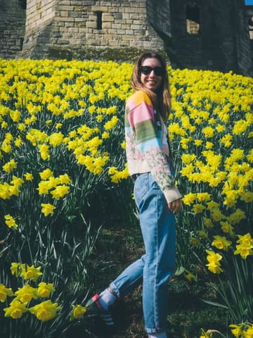 A bright, well-composed lifestyle portrait featuring a person standing in a field of daffodils with Warkworth Castle in the background. The image captures a clear, sunny day, effectively conveying a sense of seasonal travel and leisure.