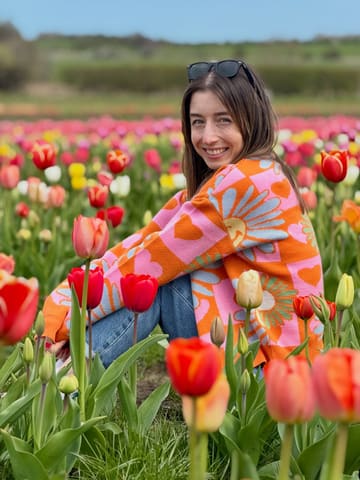 A bright, well-composed portrait of a woman sitting in a tulip field, used to promote a seasonal farm activity.