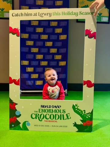 A photo of a baby positioned behind a branded photo frame prop for 'The Enormous Crocodile' musical at The Lowry. The image is a standard venue-based photo opportunity.