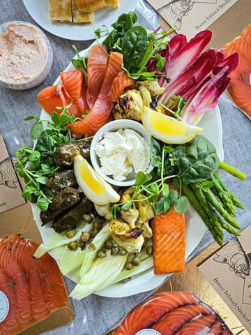A high-quality, overhead flat-lay shot of an artisanal smoked salmon platter featuring various accompaniments like artichokes, asparagus, and pate, showcasing the product's fresh and vibrant presentation.