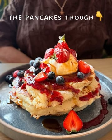A close-up, appetizing shot of a stack of pancakes topped with fresh strawberries, blueberries, and cream, served at a cafe.