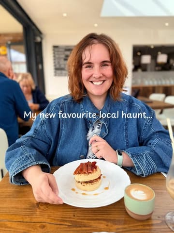 A casual, user-generated style photo of a woman at a cafe, featuring a plate of crumpets and a latte. The lighting is backlit, causing the subject's face to be in shadow.