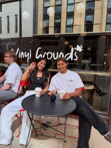A lifestyle shot of two people sitting at an outdoor table at Vinyl Grounds, holding coffee cups, with the venue's branding visible on the window behind them.