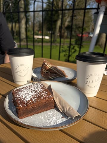 A casual, outdoor shot of a brownie, a slice of cake, and two takeaway coffee cups on a wooden table at a cafe, likely taken during a walk.
