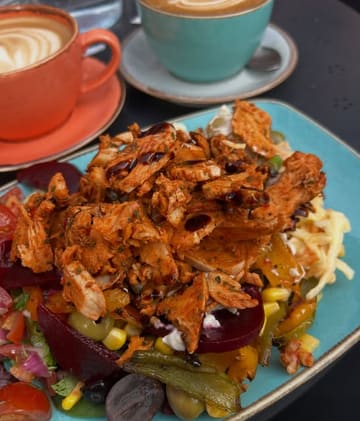 A top-down, casual shot of a chicken salad bowl served alongside two lattes at a cafe. The image captures a standard lunch setting with a focus on the main dish.