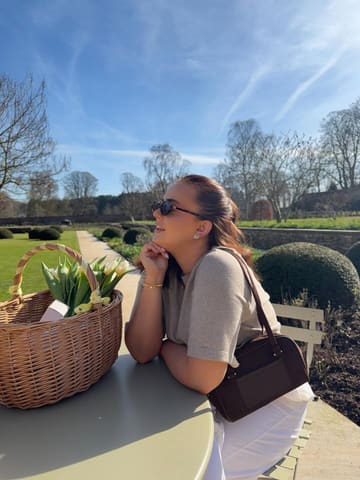 A high-quality lifestyle photograph featuring a woman sitting at an outdoor table in a garden setting at Middleton Lodge, accompanied by a basket of tulips. The image captures a bright, sunny day, conveying a relaxed and celebratory atmosphere.