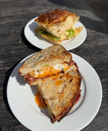 A top-down shot of two breakfast sandwiches on white plates, set against a dark wooden table. The image features a bacon and egg sandwich on sourdough in the foreground and a focaccia sandwich in the background, captured in bright, direct sunlight.