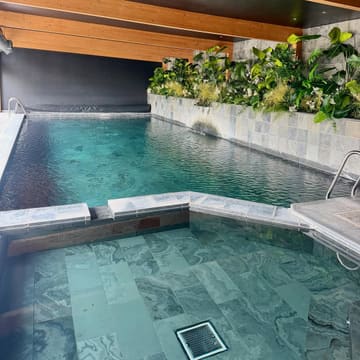 A high-quality, well-composed photograph showcasing an indoor swimming pool at a luxury accommodation venue. The image highlights the serene atmosphere, featuring clean lines, natural stone tiling, and a lush green wall, effectively communicating a relaxing and premium experience.