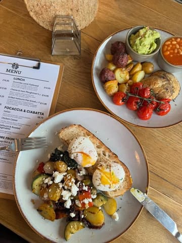 A top-down, overhead shot of two brunch plates and a menu on a wooden table at Occombe Farm. The dishes include a vegetarian breakfast with poached eggs and a plant-based breakfast with beetroot falafel.