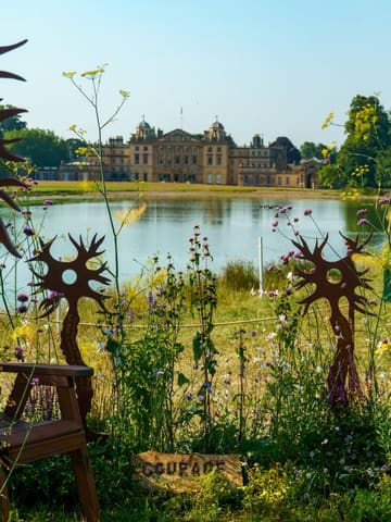 A beautifully composed, scenic shot taken at the RHS Badminton Flower Show, featuring a grand country house in the distance viewed across a lake. The image uses foreground wildflowers and artistic metal sculptures to frame the subject, creating a sense of depth and atmosphere.