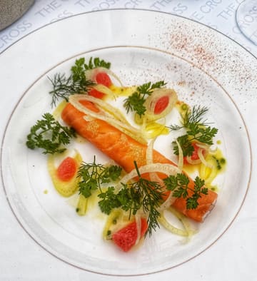 A high-quality, overhead shot of a beautifully plated salmon dish, likely a starter, served at The Cross at Kenilworth. The presentation is artistic, featuring fresh herbs, citrus segments, and shaved fennel, set against a clean, branded tablecloth.